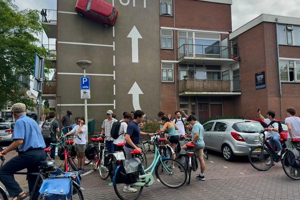 Participants pausing with their bicycles beside a facade decorated with a car mounted above painted parking markings.
