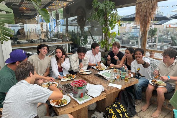 Program participants sharing a meal around a long wooden table at an open-air beach cafe.
