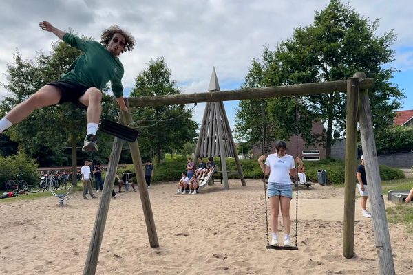 Participants playing on wooden swings in a sandy neighborhood playground, with bicycles parked nearby.