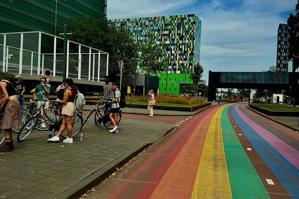 Participants standing with bicycles beside a rainbow-painted cycle street next to colorful campus buildings.