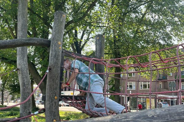 A person climbing a rope net on a wooden play structure in a leafy urban park beside a city street.