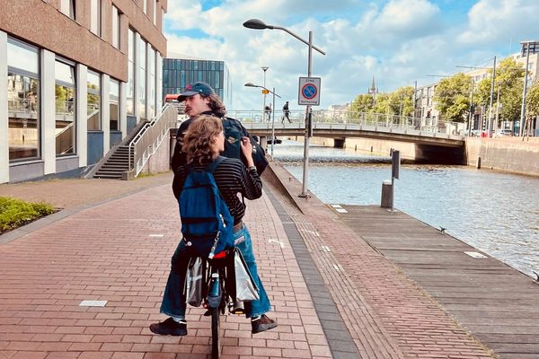 Two people riding one bicycle along a brick cycle path beside a city canal.