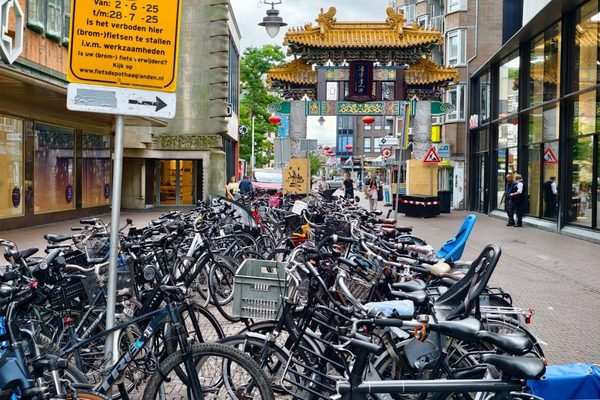 Bicycles parked densely along a city sidewalk beneath a no-parking sign, with an ornamental gateway arch behind.