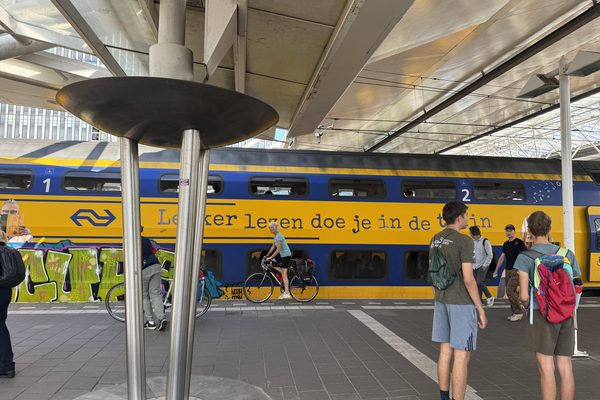 A Dutch intercity train at a covered station platform as a cyclist rides past waiting travelers.