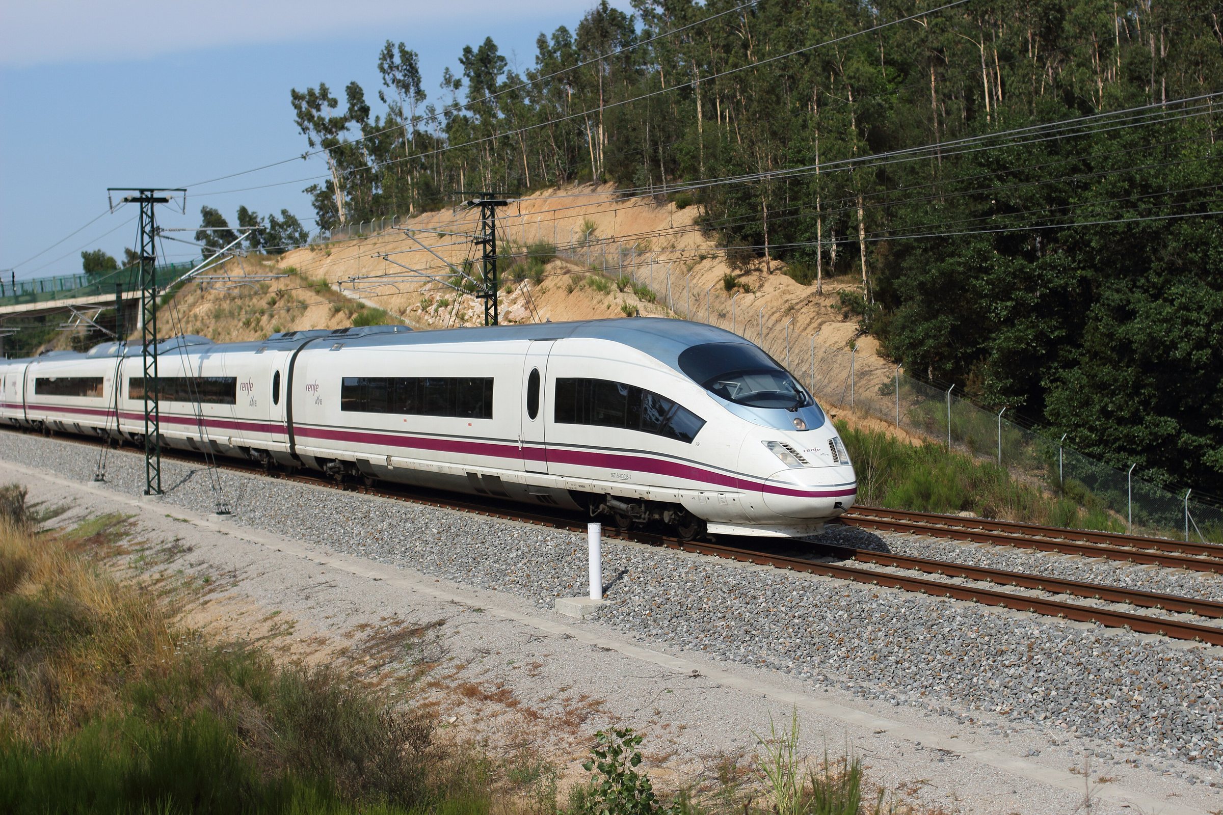 A white Renfe AVE Class 103 high-speed train on ballasted track beside a wooded hillside in Catalonia.